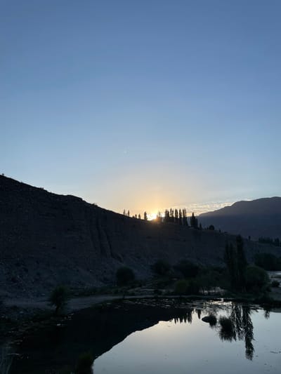 The image captures a serene twilight scene at the city of Капчыгай, Kyrgyzstan. A calm body of water occupies the lower portion of the frame, reflecting the muted colors of the sky and the surrounding landscape. The water's surface is disturbed by subtle ripples, and small clusters of vegetation are visible along its edges, also mirrored in the water.

The middle ground features a dark, textured hillside rising steeply. A line of tall, slender trees stands silhouetted against the horizon on the crest of this hill. The setting sun is partially obscured by the trees and the hillside, casting a warm, golden glow that diffuses into the otherwise clear, pale blue sky. The sky transitions to a lighter hue near the horizon, hinting at the fading daylight.

In the distance, the silhouette of rolling hills or mountains is visible, their forms softened by the twilight. The overall atmosphere is tranquil and natural, with no signs of human activity or artificial structures. The image is dominated by natural elements: water, earth, vegetation, and the sky.