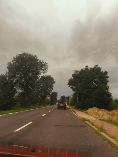 The image depicts a road scene in Olszewnica Nowa, Poland, on a cloudy day. A dark-colored car is driving on a paved road with white lane markings. To the right of the road, there is a sidewalk constructed with red and beige bricks, bordered by a curb and a patch of green grass. Beyond the sidewalk, mounds of sand and dirt are piled up. Dense green trees line the left side of the road, with one particularly large tree dominating the foreground. The sky is overcast with heavy, gray clouds. The foreground shows the orange dashboard and windshield of a vehicle.