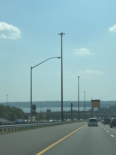 The image captures a view from a highway in Oxon Hill, Maryland, United States of America. The scene is dominated by a bright, partly cloudy sky with a few fluffy white clouds scattered across the blue expanse. A body of water, likely the Potomac River, is visible in the background, bordered by a lush, green treeline and a highway overpass structure. Several tall light poles with multiple lamps stand along the sides of the highway, illuminating the roadway. Traffic moves along the multi-lane highway, with various cars and trucks visible, including a white minivan, a dark-colored SUV, and a blue car. A yellow road sign with black lettering is mounted on a pole on the right side of the highway, indicating "ORADELL BRIDGE 1 1/4 MILES BE PREPARED TO STOP WHEN FLASHING." The road itself is paved and marked with yellow and white lane lines, suggesting normal highway conditions. The overall impression is a typical daytime scene on a busy American highway.