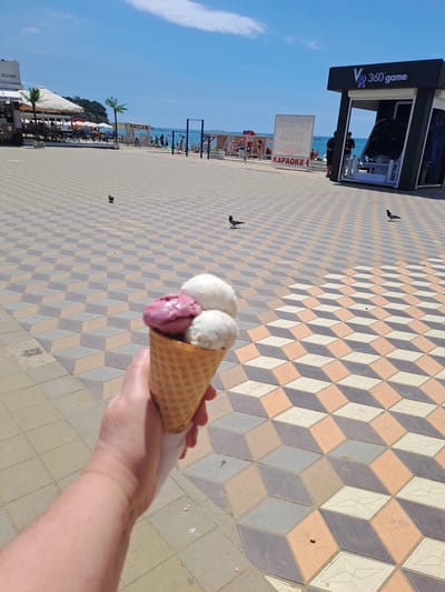 In Dzhubga, Russia, a hand holds a triple-scoop ice cream cone, featuring two white scoops and one berry-flavored scoop, in the foreground. The cone is positioned over a tiled walkway with a 3D optical illusion design of cubes in beige, grey, and white. In the background, the Black Sea coast is visible, with people walking along the beach and swimming in the water. Several buildings line the beachfront, including a cafe and a VR game booth. Pigeons are scattered across the tiled walkway. The sky is clear and blue.