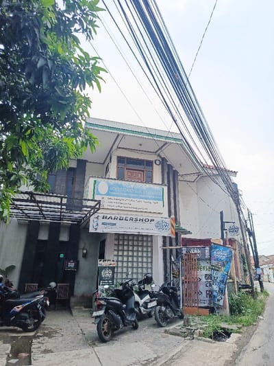 A two-story building houses a law office on the second floor and a barbershop on the ground level. Multiple scooters are parked in front of the barbershop. Overhead are numerous power lines crossing the sky. Green foliage hangs from the left. A small drain runs on the right side of the street.