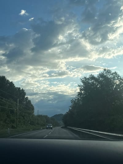The image presents a daytime view from inside a moving vehicle, looking forward along an asphalt road in a natural, mountainous setting near Bistrice, Montenegro. The sky dominates the upper portion of the frame, showcasing a mix of deep blue sky and bright, voluminous white cumulus clouds, illuminated by strong sunlight, suggesting a sunny day.
The road, with a visible white shoulder line and a partial dashed center line, extends into the distance. Lush, dense green trees and vegetation line both sides of the road, forming a thick forest that appears to cover rolling hills or the base of mountains. In the far distance, on the left side of the road, outlines of mountains are discernible under the cloudy sky.
Further down the road, a white car, possibly an SUV or van, is visible in the same lane, with its headlights on. Utility poles and power lines are also evident through the trees on the left side of the road, indicating human infrastructure in the natural landscape. The right edge of the vehicle's interior (likely the dashboard or windshield frame) occupies the foreground on the right side of the image. No visible text is present. The scene captures a moment of travel through a scenic, rural or semi-rural mountainous region.