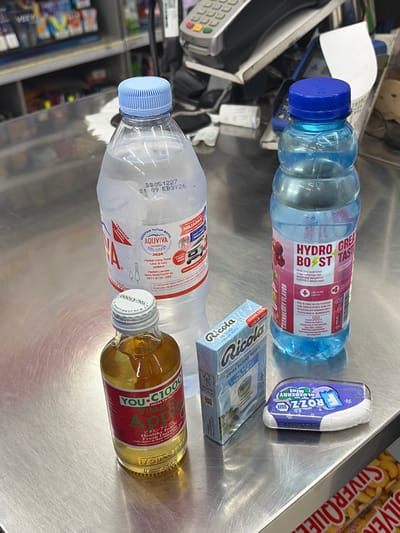 The image shows a selection of beverages and health products on a metallic counter, likely in a convenience store. Two large plastic bottles of water, one clear and one blue with a "Hydro Boost" label, are prominently displayed. To the left, a small glass bottle containing a golden liquid, labeled "YOU C1000 Vitamin Apple," is visible. A small blue box of Ricola lozenges and a white and purple package of Frozz blueberry mint pastilles are also present. In the background, blurry shelves stocked with various products and a cash register with a receipt are visible, suggesting a retail environment. The metallic surface of the counter shows some scuff marks and reflections. The presence of Indonesian labeling on some of the products and the overall context of convenience store items strongly suggests the location is Boja, Indonesia.