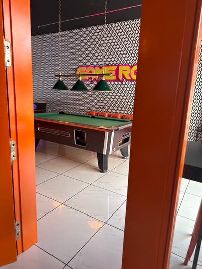 An interior scene of a game room, possibly a bar or arcade, is depicted. The focus is on a pool table with a green felt surface, a burgundy frame, and dark legs. Three green cone-shaped lamps hang above the table on a brass fixture. The background features a black ceiling and a wall covered in a repeating black and white geometric pattern wallpaper. A large, bright pink neon sign with yellow outlines spelling "GAME ROOM" is mounted on the wallpaper. To the left, an orange door with visible hinges is slightly ajar, revealing part of the room. The floor is tiled with large, white, marble-patterned tiles. Orange stools are visible along the side of the pool table. The image suggests a retro or vintage-themed establishment. There are no people visible in the scene.