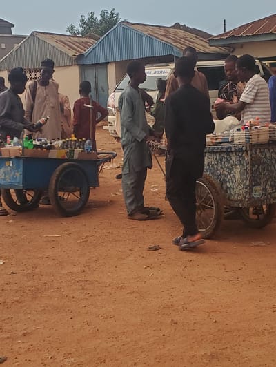 This is a media file depicting an outdoor scene in Funtua, Nigeria. The primary subjects are a group of people interacting around a mobile cart filled with various bottled drinks. The people appear to be of various ages and genders, dressed in casual clothing. Some are standing, while others are seated.
The central object is a cart, which is brightly colored and appears to be used as a vendor's stall for selling beverages. The cart has wheels, suggesting it is mobile. Behind the cart, there are buildings with corrugated iron roofs and painted walls, characteristic of structures in this region. The ground is sandy and appears dry, indicating fair weather.
The activity taking place seems to be a casual gathering or a commercial transaction, with people potentially buying or selling the drinks displayed on the cart. The atmosphere appears relaxed, with no immediate signs of urgency or distress. The time of day is not explicitly clear, but the lighting suggests it might be late afternoon.
Visible text on the cart is difficult to decipher due to the image's resolution and orientation, but a sign on the building reads "DA KYAU". This phrase is likely in Hausa, meaning "it is beautiful" or "good". The overall scene captures a slice of everyday life in a Nigerian town.