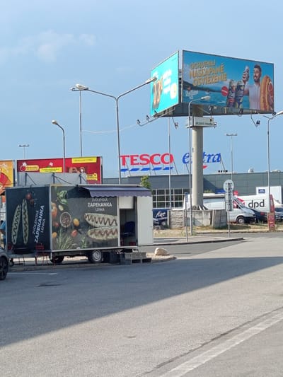 This media file is a photograph taken outdoors, likely during the daytime with clear skies, as indicated by the bright sunlight and shadows. The scene is dominated by a street and several commercial establishments, suggesting an urban or suburban setting.
In the foreground, a food truck is prominently featured, displaying large advertisements for "ZVOLENSKÁ ZAPEKANKA LENKA." These advertisements include appetizing images of the food item.
Further back, a large "TESCO" supermarket sign is visible, as well as a sign for "Biedronka," another grocery store. Adjacent to Tesco is a building with "tra" logo visible on it, and another building with "dpd" logo on it. This indicates a commercial area with multiple retail outlets.
Several billboards are also present. One billboard promotes a beverage with the text "OCHUTNAJ NASLAPANE OSVIEZENIE" and images of cans and a person holding a ring. Another billboard displays a sale or promotion, with text including "OD 25. 6. DO 1/7.2026" and "CENA."
The overall impression is a bustling commercial zone, possibly a shopping center or a street with various businesses. The presence of streetlights and the angle of the sun suggest it is likely daytime. There are no explicit people featured prominently, but the context points to a public, commercial space. The text is primarily in Slovak, further reinforcing the location context of Zvolen, Slovakia.