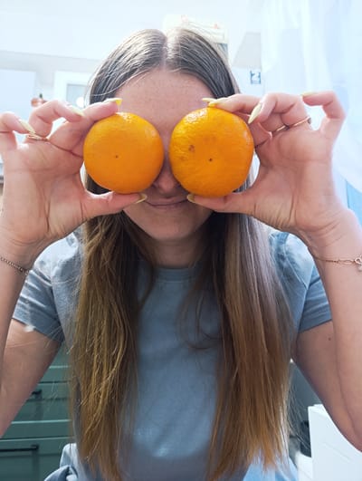 A young woman holds two oranges in front of her eyes. Her long, brown hair hangs down her shoulders. She wears a blue t-shirt, gold rings, and bracelets. The background shows a room with cabinets and a window with sheer curtains. There is no evidence to suggest a location in the city of Chernogolovka, Russia.