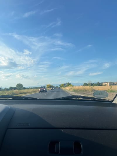 This is a first-person perspective image taken from inside a vehicle traveling on a road in Graf Ignatievo, Bulgaria. The dashboard is visible in the foreground, showing the "AIRBAG" logo. The road ahead is lined with dry grass and vegetation, with trees and buildings visible in the distance on either side. Several vehicles are on the road, including a red SUV in the middle distance. The sky is a clear blue with wispy white clouds. The image appears to have been taken during daylight.