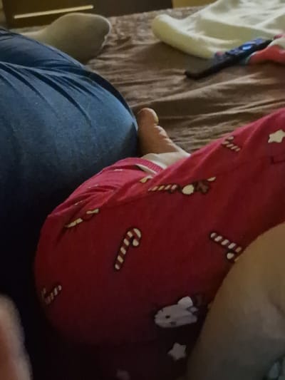 The image shows a person wearing blue jeans and red patterned pajama pants with candy cane, reindeer, and star designs. The person is resting their bare foot on their leg, and there is bedding visible in the background. The image provides no information regarding location in Byala Slatina, Bulgaria.