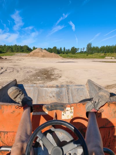 The image is a first-person perspective from the cab of an orange construction vehicle, likely a front loader. The operator's legs and feet, clad in dark work boots, are visible in the foreground, positioned above a steering wheel. The vehicle's bucket, appearing worn and metallic, is in front of the operator. The scene outside the vehicle depicts a sandy, open area with a large pile of light-brown material, presumably aggregate or sand, in the mid-ground. A line of green trees forms the background, under a bright blue sky with wispy white clouds. The overall impression is that of an outdoor work site, possibly a construction or quarrying location. The specific location in Skrunda-1, Latvia, is not discernible from the visual cues in the image itself.