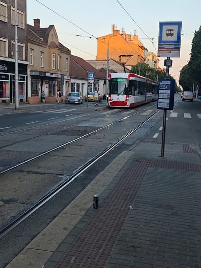 This is a medium-shot, eye-level photograph taken on a street in Brno, Czechia. The scene captures a tram with the number 1856 on its front, adorned in red and white, traveling down tram tracks. The tram is illuminated by its headlights and displays "1 NECHAL" on its digital sign. In the background, buildings line the street, some with storefronts, including one labeled "SPORT." A blue tram stop sign with pictograms and text like "JUNGMANNOVA" and route information is visible. Several people are walking on the sidewalk and crossing the street. The time of day appears to be late afternoon or early evening, with a warm, golden light casting long shadows, suggesting a clear sky. The setting is urban, with paved sidewalks, tram tracks, and overhead wires for the tram system. A discarded can of soda rests on the curb near the tram tracks. The overall mood is one of everyday urban life.