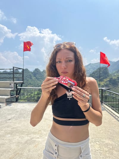 A young woman stands on a concrete surface in Lũng Phìn Commune, Vietnam, against a backdrop of mountains and a partly cloudy sky. She is holding a Kit Kat Chunky candy bar. Two red flags are visible.