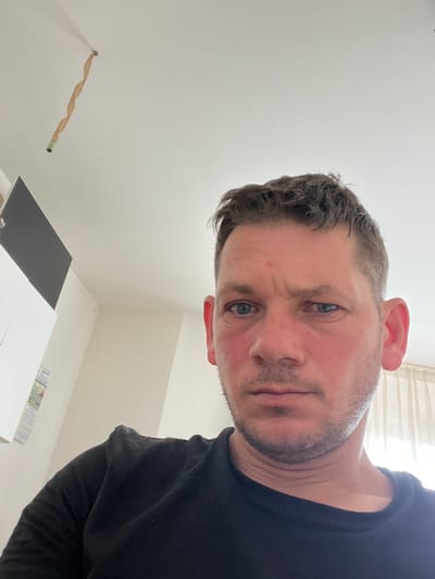 The image is a selfie taken indoors, likely in an apartment. The subject is a white male with short, dark hair, blue eyes, and a fair complexion. He is wearing a black t-shirt. His face is slightly flushed, and he has a concerned or questioning expression.
In the background, a white ceiling is visible, with a twisted paper decoration hanging from it, attached to a small green object at the bottom. To the left, there are two white wall-mounted units, one with a dark gray rectangular panel on top, and a white one below with some papers attached. The right side of the image shows a cream-colored curtain.
The specific location in Senago, Italy, cannot be determined from the visual information provided in the image.