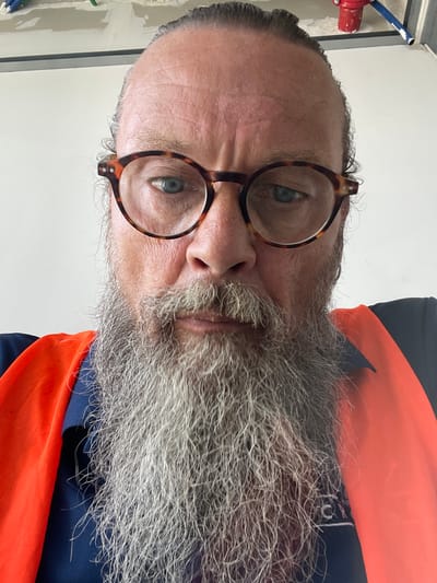 A close-up, overhead shot shows a man with a large, gray beard and tortoiseshell glasses. He is wearing a navy blue shirt and an orange safety vest. The background appears to be an indoor construction site in Ramot HaShavim, Israel, with exposed wiring and pipes visible on a light-colored wall. The man is looking directly at the camera.