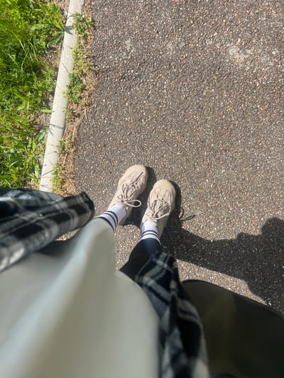 An elevated view depicts a person standing on a gray asphalt surface with two light brown sneakers with light laces. They are wearing white socks with black stripes at the top and dark-colored pants underneath a light colored, possibly white, top. A black and white checkered garment is draped across the lower right quadrant of the view. The edge of a concrete curb and green grass are visible at the bottom left corner. A shadow cast from the person/object can be observed on the asphalt surface. The location cannot be determined from the image.