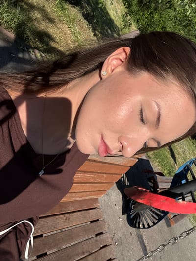 The image shows a close-up of a young woman sitting outdoors, with her eyes closed. She has dark brown hair and is wearing a brown top. A delicate gold necklace with a small white charm rests on her collarbone. Her skin appears smooth and slightly luminous, catching the sunlight. She has a subtle earring in her left ear, a small, delicate stud with a floral design.

Behind her, the scene suggests a park or outdoor recreational area. To the left, green grass and trees are visible, dappled with sunlight and shadows, indicating it is daytime. To the right, a wooden bench is partially visible, and beyond that, what appears to be a playground structure with a red and black propeller-like element and metal chains. The ground is a mix of concrete and wood.

The overall impression is one of peacefulness and relaxation, with the woman enjoying the warmth of the sun. The lighting suggests it is a sunny day, likely late morning or afternoon given the angle of the shadows. The presence of playground equipment and the natural setting place the location in an outdoor, public space, possibly a park in Belovo, Russia. There is no visible text in the image.