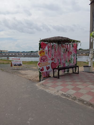 The image shows an outdoor scene in Sol-Iletsk, Russia, likely near a recreational area. In the foreground, a paved walkway with a checkered pattern of pink and gray tiles leads towards a decorated structure. This structure appears to be a photo booth or a decorative alcove, with a thatched roof and walls draped with cascading artificial pink and white flowers. A dark wooden bench sits in front of the flower-covered wall. To the left of the structure, a sign with red text on a white background and an arrow indicates an attraction, with an image of ATVs and text stating "for children and adults." Further in the background, a body of water, possibly a lake or pool, is visible with a long, white fence running along its edge. Beyond the water, there are buildings and a cloudy sky. A utility pole with wires stands to the right of the flower-draped structure.