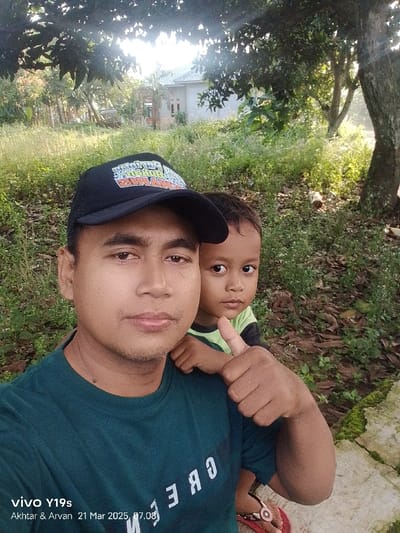 The image shows an adult male and a young boy standing in an outdoor setting. The adult male is wearing a black baseball cap with white lettering, a green t-shirt, and has short dark hair. The young boy is wearing a green t-shirt and has dark hair. The adult male has his right arm around the young boy's shoulders and his right thumb is pointed upward. The young boy is facing the camera and smiling. The background includes green plants, a partially visible house, and a large tree. The image is taken in a location in the city of Kecamatan Anjatan, Indonesia, with coordinates -6.417 latitude and 107.978 longitude. The ground is covered in dry leaves and green grass. There is a concrete walkway on the right side of the image. The adult male's left hand is not visible. The image is a close-up shot taken with a smartphone. The time stamp on the photo is 21 Mar 2025, 07:08. The phone model is vivo Y19s. The image was taken by Akhtar & Arvan.