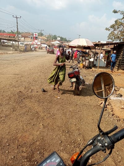 This image depicts a bustling street scene in Aramoko, Nigeria. The foreground shows the handlebars, speedometer (reading 0 km/h), and right rearview mirror of a motorcycle, indicating the perspective of the photographer. The ground is an unpaved, reddish-brown dirt surface, dusty and uneven. A dark-colored pigeon stands on the dirt road, near a small, light-colored rectangular object.
In the midground, a slender individual, dressed in a loose olive-green jumpsuit and flip-flops, walks towards the right side of the frame, holding a light-colored object in their right hand. Directly behind them is a parked motorcycle, viewed from the rear, displaying a Nigerian license plate "KDM 522 QA." To the right, a market area features several open stalls with corrugated metal roofs and a prominent light-pink parasol shading goods and vendors. Multiple individuals are visible near these stalls, some wearing red garments, and a man in a blue shirt and jeans stands beside a wooden structure.
The background reveals more of the unpaved road, along which a red sedan drives away from the camera. On the left side of the road, a "Total" petrol station sign, red and white, is visible with fuel price indicators. Various single and multi-story buildings with reddish roofs line the street, connected by numerous utility poles and overhead power lines. The sky is partly cloudy, with patches of clear blue.
