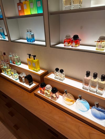 The image shows a display of perfumes in a store, likely a high-end department store or boutique. The store appears to be located in Xuanwu District, China. The display features multiple shelves with various perfume bottles arranged in an organized and aesthetically pleasing manner. The lighting is bright, highlighting the colors and shapes of the bottles. Some of the perfumes are in clear glass bottles, showcasing their liquid contents which range in color from clear to yellow and blue. Other bottles are opaque or have colored labels. The display also includes some decorative elements, such as ribbons and caps, that add to the overall presentation. The background is neutral, with textured walls and wooden shelves, drawing attention to the products themselves. There are no people visible in the image. The scene is clean and well-maintained, suggesting a professional retail environment.