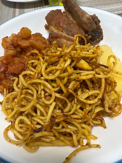A close-up image presents a meal served on a white oval plate featuring a thin blue rim. The plate holds four distinct food components. In the upper right quadrant, a single piece of dark brown, roasted or fried meat, appearing to be a bone-in cut, exhibits a visibly crisp exterior. Dominating the center and lower right, a large quantity of pan-fried noodles, resembling spaghetti, is arranged. These noodles are golden-brown with areas of darker, crispy texture, and incorporate small, coiled pieces, possibly of fried onion. To the far right, next to the noodles, are several irregular, pale yellow, somewhat translucent pieces, consistent with boiled yuca or potato slices. In the upper left quadrant, a reddish-brown stew or saucy dish is visible, containing numerous small, irregularly cut pieces of a starchy vegetable, likely potatoes, within a thick, glossy sauce. The background is blurred, showing a patterned grey and white textile surface on the left and a partial view of a yellow-green object in the upper distance. The meal is depicted as being located in Santiago de Surco, Peru.