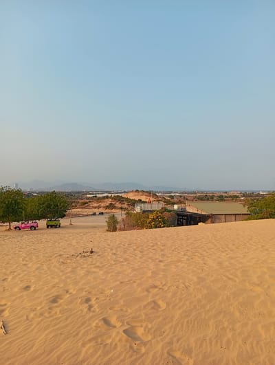 The image depicts a sandy landscape, possibly a sand dune, in Phuoc Dinh, Vietnam. The foreground consists of sand with wind-blown patterns. In the mid-ground, there is some vegetation and a building with a corrugated metal roof. Two open-top vehicles, one pink and one green, are parked under trees. In the background, a cityscape is visible with mountains in the distance under a clear sky.