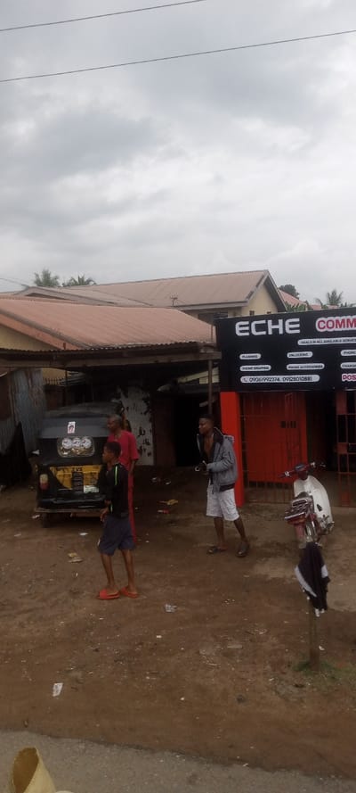 This image depicts an outdoor urban scene in Ezinifite, Nigeria, under an overcast sky. The foreground shows a bare reddish-brown dirt ground with scattered debris, transitioning to a narrow strip of paved road at the bottom. Three males are present: a young male in a black shirt, dark shorts, and red sandals stands foreground-left, facing left; an older male in a red top and dark trousers stands midground-left behind a parked black and yellow Bajaj RE auto-rickshaw; and a young male in a gray hooded jacket, white distressed shorts, and dark sandals stands midground-right, holding a dark object in his left hand, facing left.
The background features multiple structures. On the left, a building with a brown corrugated metal roof and corrugated metal fencing is visible. To the center-right, a black signboard for "ECHE COMMUNICATION" advertises services including "FLASHING," "UNLOCKING," "PHONE SWAPPING," "PHONE ACCESSORIES," "CHARGER," and "POS," along with two phone numbers (09061992374, 09128010881). Below the sign are closed red metal roll-down security doors. To the right of the signboard, a white Honda Cub-style motorcycle with red accents is parked, facing left, with a black garment draped over an adjacent pole. Further in the background, more buildings with brown corrugated roofs and scattered palm foliage are discernible. Two overhead power lines traverse the upper portion of the frame.