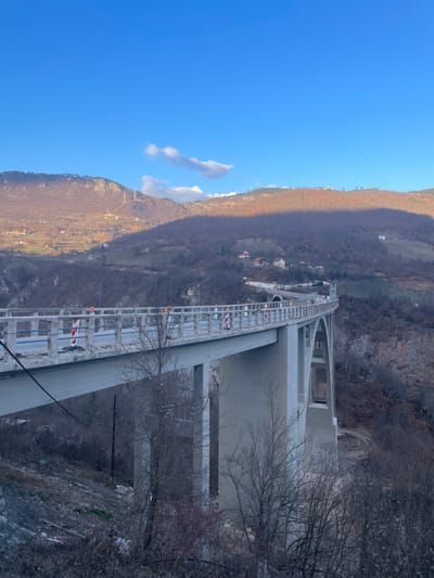 This image depicts the Đurđevića Tara Bridge, located in Đurđevića Tara, Montenegro. The scene is an elevated, panoramic view of the multi-arched concrete bridge spanning a deep, forested canyon under a clear, bright blue sky with scattered white clouds.
The bridge, constructed of light grey concrete, features a roadway marked by numerous orange and white striped traffic cones or bollards, indicating ongoing maintenance or construction. Several individuals, identifiable as workers by their orange high-visibility clothing, are present on the bridge deck. Blue scaffolding or netting is visible along the inner edge of the bridge's railing. The bridge's substantial concrete piers and arches descend into the valley below.
The surrounding environment consists of steep, mountainous terrain, predominantly covered with bare deciduous trees, suggesting a late autumn or winter season. The left mountain slope is brightly illuminated by sunlight, revealing scattered houses and utility poles. In contrast, the right mountain slope is largely enveloped in shadow. The immediate foreground features a sparse arrangement of bare tree branches. The bottom of the canyon is not directly visible from this perspective.