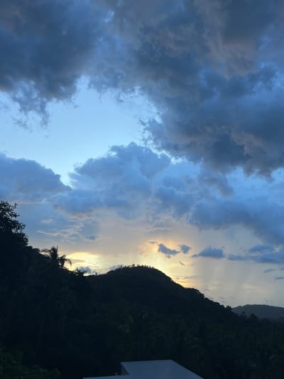The image captures a dramatic sunset over a lush, mountainous landscape in Baan Harn, Thailand. The sky is a symphony of blues and grays, with dark, heavy clouds dominating the left side of the frame, gradually giving way to lighter, wispy clouds and shafts of golden sunlight on the right. These sunbeams break through the atmospheric veil, creating a celestial spectacle.
The foreground is dominated by the silhouette of a dense, dark forest covering the steep slopes of the mountains. The foliage is thick and indistinguishable in the low light, with hints of palm trees visible. The top edge of the frame reveals a portion of a white building, suggesting a structure or resort at a higher elevation, providing a vantage point for this view.
The time of day is clearly sunset, indicated by the warm, golden light emanating from the horizon and the dramatic cloud formations. The weather appears to be transitional, with the dark clouds suggesting a recent or impending rain shower, juxtaposed with the brilliant sunshine breaking through. The overall mood is one of awe and serenity, with the natural beauty of the landscape illuminated by the fading light of day. No text is visible in the image.