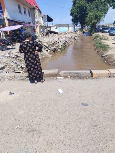 The image depicts a street scene in Tursunzoda, Tajikistan, characterized by a central drainage ditch filled with murky brown water. The ditch is flanked by uneven, debris-strewn banks, with rubble and various discarded items visible along its edges. On the left side of the ditch, a partially demolished building stands, showing signs of severe damage and disrepair. In front of this structure, a market stall with purple awnings is set up, displaying goods such as shoes and clothing. A person wearing a floral-patterned dress and a headscarf stands facing away from the camera, near the edge of the ditch. Further down the street, on the right side of the ditch, a line of trees shades the sidewalk where several parked cars are visible. The overall environment appears to be a mix of urban decay and everyday commerce.