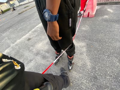 A high-angle shot shows two people standing on an asphalt surface. One individual wears inline skates and black trousers. A red and white tape is stretched between their legs. The other person wears black trousers and black shoes. The surface appears to be part of a paved area, possibly a parking lot or street. A building is visible in the background. There is no indication of the location being in Sangotedo, Nigeria, in the image. No interaction is occurring between the two people. The time of day is daytime, evidenced by the clear visibility.