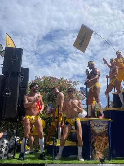 The image depicts a vibrant scene in Sitges, Spain, likely during a parade or festival. Several men, mostly shirtless and wearing yellow swim briefs adorned with soccer ball motifs, are standing on a decorated platform. Some are holding drinks, flags, or fans. A large yellow flag with the "Stay Sitges Accommodation" logo is prominently displayed. The background features lush green foliage and a partly cloudy sky. Large speakers are visible to the left, and a colorful mural with soccer-themed imagery and text like "ONE WORLD ONE LOVE" is on the side of the platform. The overall atmosphere suggests a celebratory and lively event.