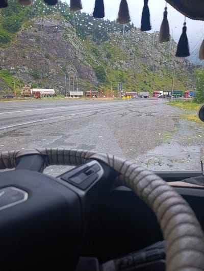 The image shows a view from inside a truck cab, looking out at a border crossing or checkpoint in Nizhnii Lars, Russia. The foreground is dominated by the steering wheel of the truck, which is wrapped in a light-colored fabric. Beyond the steering wheel, the road is wet and appears to have puddles, suggesting recent rain. A mountainous, rocky terrain with sparse vegetation rises in the background. Several trucks are visible at the checkpoint, some lined up, indicating traffic. There are also buildings and structures associated with a border control area, including what appears to be a toll booth or barrier system. Flags are flying on poles. The sky is overcast, contributing to the damp atmosphere. The scene captures a moment at a busy transportation hub in a mountainous region.