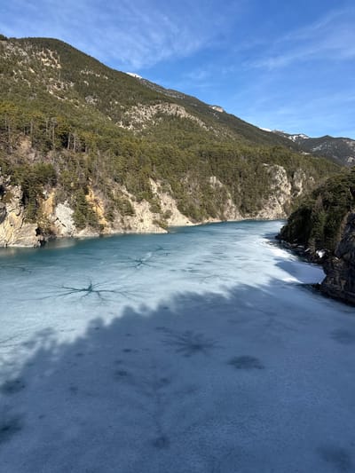 The image displays a panoramic view of a partially frozen segment of the Durance River, or a connected reservoir, located within a mountainous valley near Briançon, France. The central and lower portions of the frame are dominated by the body of water, which exhibits a distinct turquoise coloration. A substantial area of this water is covered by light blue ice. The ice surface features multiple dark, radial fracture patterns resembling starbursts, prominently visible in the mid-ground. A large, irregular dark shadow, originating from an unseen source, extends across the foreground and mid-ground of the frozen surface. Sections of open, unfrozen turquoise water are observable further upstream towards the left.

The river is bordered by steep, rugged mountainsides rising directly from the water's edge. The left bank exposes prominent light-colored rock cliffs, partially obscured by sections of dark green coniferous forest that densely cover the upper slopes. Similar forested terrain lines the right bank. In the background, higher mountain peaks are visible, displaying scattered snow cover. The sky above is a clear, vibrant blue with minimal wispy white clouds. The scene is illuminated by direct sunlight, casting sharp shadows. No individuals, animals, or specific man-made objects are identifiable within the frame.