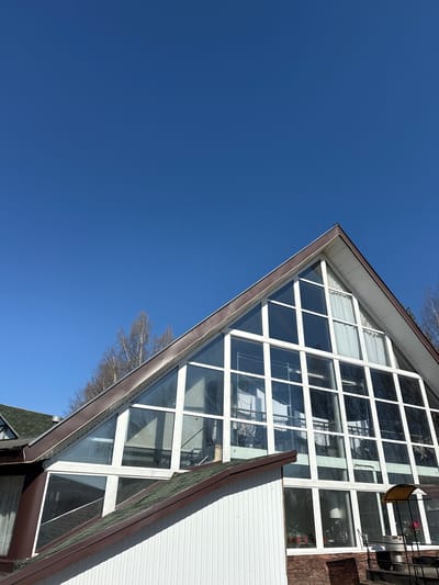 A low-angle shot shows a building facade against a clear blue sky. The building has a large, triangular window section with a grid of white frames. A balcony with a chair and some plants can be seen through the windows. The facade appears to be partly brick and partly white siding. Some bare tree branches are visible in the upper left of the frame. Given the prompt context, this image may be taken in Галево, Russia.