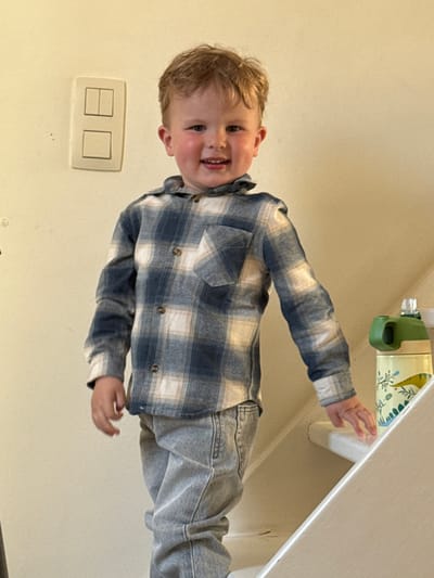 This indoor portrait features a cheerful young child, likely a toddler or preschooler, captured in Bredene, Belgium. The child has light brown, wavy hair and fair skin, and is looking directly at the camera with a wide, open-mouthed smile, revealing their teeth. They are dressed in a long-sleeved plaid shirt featuring blue and white squares, paired with light-colored denim jeans.
The child's right hand (viewer's left) is slightly raised, while their left hand (viewer's right) rests on or reaches towards a white surface, possibly a counter or shelf. On this surface, a patterned object, which appears to be a water bottle, is visible with a green handle and a design featuring yellow and blue-green elements.
The background is simple, consisting of plain, light-colored walls, likely beige or cream. On the wall to the child's left, a light switch plate with two rectangular switches is visible; one switch appears to be in an 'on' position. The lighting is soft and even, typical of an indoor setting during the day. The overall impression is one of happiness and childlike innocence. There is no visible text in the image.