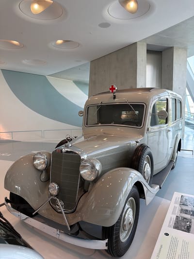 This is a full shot of a vintage Mercedes-Benz ambulance, likely on display in the Mercedes-Benz Museum in Stuttgart, Germany. The ambulance is a light beige color with chrome accents. It features a prominent grille, large headlights, and a spare tire mounted on the side. A red cross emblem is visible on the roof, signifying its purpose. The interior of the vehicle is visible through the windows. The ambulance is positioned on a white platform, suggesting a museum or exhibition setting. In the background, there is a modern architectural design with concrete walls and ceiling lights, consistent with the Mercedes-Benz Museum's aesthetic. The lighting is bright and diffused, illuminating the vehicle and its surroundings. There are no people visible in the image. The scene is clean and well-maintained, typical of a museum environment.