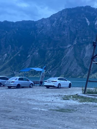 The image depicts a serene, overcast scene at a lakeside location, potentially in the vicinity of Irganay, Russia, given the characteristic mountainous terrain and water body. In the foreground, a gravelly shore is visible, with three cars parked: a silver sedan on the left, a white sedan in the center, and another white sedan partially visible on the right. Adjacent to the center car is a blue canopy structure providing shade, under which a wooden bench is placed. Two surfboards are leaning upright near the canopy. The middle ground features a calm body of water, likely a lake or reservoir, with the vast, rugged mountains forming the backdrop. The mountains are characterized by steep slopes, rocky surfaces, and sparse vegetation. The sky is uniformly gray with cloud cover, contributing to the subdued lighting of the scene. No individuals are present in the image.