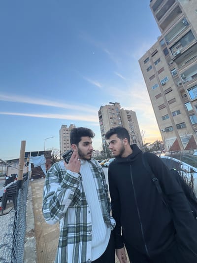Two men stand outdoors with buildings in the background and a bright sky. The man closest to the viewer wears a white sweater with a green and white plaid shirt over it and is on a phone call. The other man, behind him, has a black jacket with a black shoulder bag. Both men have dark hair. A fence and other objects are in the foreground.