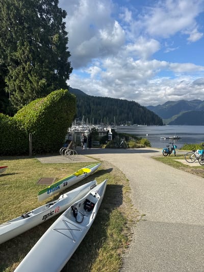 This image depicts a waterfront scene in North Vancouver, Canada, likely at a park or marina. In the foreground, three kayaks are resting on a grassy area next to a paved pathway. The kayaks are white and one has green and yellow accents with the number "67" visible. Further down the path, several bicycles are parked.
In the middle ground, a well-manicured hedge shaped like a large, abstract sculpture stands prominently on the left. Behind the hedge, a few people are gathered near a bike rack and a sign. A marina with numerous sailboats and yachts is visible in the background, stretching across a calm body of water.
The background is dominated by a tree-covered mountainside, which recedes into the distance under a partly cloudy sky. The overall impression is of a serene and recreational outdoor setting.