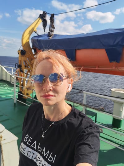 In Veregonets, Russia, a woman with blonde hair, wearing sunglasses and a black t-shirt with text that appears to be in Russian, stands on the deck of a vessel. The t-shirt has the words "ВЕДЬ МЫ НЕ СТАРЕЮ" printed on it. She is wearing a silver necklace with a small pendant. White wireless earbuds are visible in her ears. In the background, there is a yellow crane, an orange and blue lifeboat, and the water. The sky is partly cloudy. The deck is green, and there is a metal railing. The overall scene suggests a day trip or voyage.