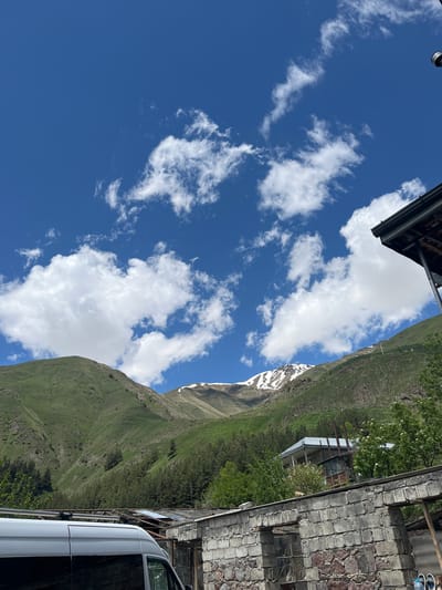 This media file captures a scenic view of a mountainous landscape in Tkarsheti, Georgia. The scene is dominated by a vast, clear blue sky filled with scattered white cumulus clouds. In the foreground, on the right side of the image, a white vehicle is partially visible, parked next to a building constructed with a combination of stone and brick. The building appears rustic, with a corrugated metal roof and exposed structural elements. Beyond the building, the lush green slopes of a mountain rise steeply. Patches of snow are visible on the higher elevations of the mountain, suggesting a high-altitude or early-summer setting. Trees with vibrant green foliage dot the mountainside. The overall impression is one of natural beauty and a rural, mountainous environment. The lighting suggests it is daytime, likely late morning or early afternoon, with bright sunlight illuminating the scene. There are no visible people or text in the image.