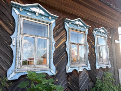 The image displays the exterior of a weathered wooden house in Niva, Russia. Three intricately decorated windows, framed by blue wooden trim with white detailing, are visible. The window frames are adorned with carvings of birds and geometric patterns, showcasing traditional Russian wooden architecture. The wooden siding of the house is dark and appears aged, with diagonal planks. Green foliage is growing around the base of the house, partially obscuring the lower portions of the windows. The windows reflect the sky and surrounding environment, suggesting it is daytime. A street sign with Cyrillic lettering is partially visible on the right side of the image. There are no people present in the scene.
