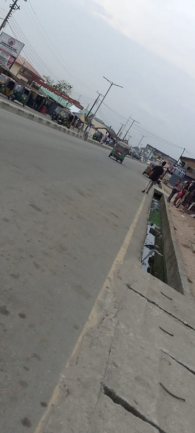 The image captures a street scene from a slightly elevated and tilted perspective, looking down a road that stretches into the distance under an overcast sky.
In the immediate foreground, a concrete drainage ditch runs parallel to the road on the right. The ditch contains murky, greenish water and some visible debris. The concrete structure shows signs of wear and cracking. Adjacent to the ditch, on the left, is an asphalt road with a faded yellow line marking its edge. The road surface has several dark stains or patches.
On the left side of the road in the midground, numerous three-wheeled vehicles, commonly known as tricycles or "keke Napeps," are parked or waiting. Behind these vehicles, there are various roadside stalls or shops with corrugated metal roofs and awnings, indicating a commercial area. Above these structures, a large white billboard is prominently displayed. The legible text on this billboard reads: "MOUNTAIN OF FIRE AND MIRACLES MINISTRIES." Below this main text, smaller lines of text are visible but too indistinct to read, likely detailing service information or church activities. Another sign below it has red lettering, with "INVESTMENT LTD" clearly visible, though the preceding word is unclear.
On the right side of the road, further down from the foreground ditch, a person wearing a dark top and light trousers is walking or standing near the edge. Other individuals are also visible along the roadside, near what appears to be a dirt or unpaved shoulder beyond the concrete ditch. Another tricycle is seen driving away in the distance on the right side of the road.
The background features more buildings, characteristic of an urban or semi-urban environment. Numerous utility poles with a network of overhead wires crisscross the upper part of the image against the light grey, cloudy sky. Modern streetlights with T-shaped fixtures are also visible along the street.
The overall impression is that of a bustling street during the daytime, likely an overcast day given the diffuse lighting, with commercial activity and public transportation present.
