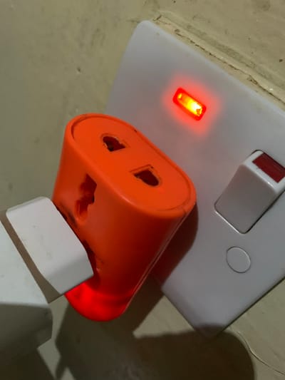 This media file shows a close-up view of an electrical outlet in Nabatum, Nigeria. The primary subjects are an electrical socket with a red indicator light, a bright orange universal plug adapter, and a white plug, likely from a device. The socket is mounted on a white wall plate, and a light switch is visible at the top of the plate. The orange adapter is plugged into the socket, and the white plug is inserted into the adapter. A red light is illuminated on the adapter, indicating that it is receiving power.
The scene is lit by artificial light, as suggested by the illuminated red indicator lights and the shadows cast on the wall. The wall itself appears to be textured and light-colored, with some visible imperfections. The overall impression is of an indoor electrical setup. No specific time of day or weather conditions can be inferred from the image. No people are visible. The activity taking place is the charging or powering of an electrical device via the adapter. The image focuses on the details of the electrical connections, highlighting the functional aspect of the scene. No text is visible in the image.