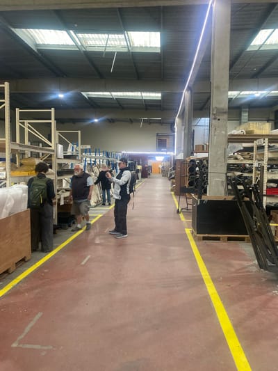 An indoor scene depicts a spacious industrial warehouse or workshop, likely in Montreuil, France. The primary subjects are four individuals, all appearing to be adult males, engaged in observing or inspecting materials stored on tall metal shelving units.
The floor is a reddish-brown concrete, marked with a prominent yellow safety line that delineates a walkway down the center of the facility. Along both sides of this aisle, multiple industrial-style metal racks are filled with various raw materials and components, including rolls of fabric or insulation, metal bars, and wooden planks. Visible text on the shelves includes "METAL", "BOIS" (French for wood), and partially visible "LUX" and "RS". Wooden pallets, some marked with "EUR", are also present.
The ceiling is high, featuring exposed concrete beams and corrugated metal panels. Bright strip lighting runs along the ceiling, complementing the natural light streaming in through large, high-set windows.
One person, wearing a black and white jacket, dark pants, and a cap, with a backpack, stands in the foreground, looking downwards. Further down the aisle, another individual with a white beard is seen in a dark vest and shorts, also appearing focused on items on the shelves. A third person, with dark curly hair and a green backpack, stands with their back to the viewer, near the shelves. A fourth person is partially visible behind the first individual. The activity suggests an inspection, inventory, or a guided tour of the facility. The lighting indicates it is daytime.