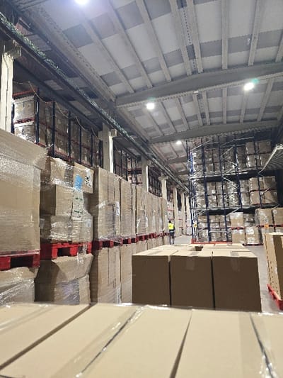 The image depicts the interior of a large industrial warehouse located in Valverde del Majano, Spain. The scene is dominated by extensive storage of goods. In the foreground, several rows of unsealed, standard brown cardboard boxes are present, resting on what appears to be the concrete floor. Behind these, numerous stacks of shrink-wrapped cardboard boxes are piled high, supported by red wooden pallets. These stacks form a continuous wall along the left side of the frame and extend into the mid-ground. Further into the background and on the right, a multi-tiered metal racking system is visible, also densely packed with similar palletized, shrink-wrapped boxes.
The warehouse features a high, industrial-style ceiling composed of corrugated panels and structural beams, from which multiple circular, bright artificial light fixtures are suspended, providing illumination. The floor is a flat, untextured concrete surface.
One individual is visible in the mid-ground, positioned between the stacked goods. This person is wearing a high-visibility yellow or lime-green vest over dark attire and appears to be walking away from the camera, deeper into the warehouse aisle. No direct interaction between the individual and the objects is discernible. The overall environment suggests active, large-scale storage and logistics operations.