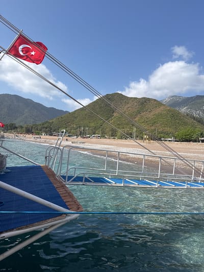 The image captures a view from a boat near the shoreline in Kumluca, Turkey. A sandy beach runs parallel to the mountainous terrain visible in the background. The water near the boat is clear and turquoise. On the boat itself, a Turkish flag is attached to one of the ropes. The boat also has a ladder with blue steps leading down to the water. The scene is set under a blue sky with scattered clouds. The beach is lined with trees and other structures. People are visible on the beach.