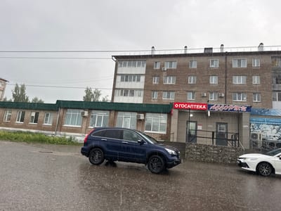 This image depicts a rainy, overcast day on a street in Mozhga, Russia. The scene is dominated by wet asphalt, showing numerous visible raindrops creating splashes and reflections on the ground, indicative of heavy rainfall.

In the foreground, a dark blue SUV, possibly a Honda CR-V, is positioned on the wet road, its dark wheels prominent against the grey surface. To its right, the front section of a white sedan, resembling a Toyota Camry, is parked.

The midground features two buildings. On the left, a single-story brick building with a green roof houses commercial spaces. Behind it, a multi-story brick apartment building extends across the frame, characterized by numerous windows and several external air conditioning units. Overhead, several power lines stretch horizontally across the dull, grey sky. Trees are visible in the background on the left side.

Visible text on the commercial signs includes "ГОСАПТЕКА" (GOSAPTEKA), which translates to "State Pharmacy," displayed in red letters. Another prominent sign reads "АССОРТИ" (ASSORTI), meaning "Assortment," in blue. Smaller signs mention "Товары для пикника" (Goods for picnic) and "Домашняя выпечка" (Homemade baked goods), along with a "ZANUSSI" logo.

The time of day appears to be daytime, though the heavy cloud cover gives a subdued light. No people are visible in the image. The overall mood conveyed is one of a typical rainy day in an urban setting.