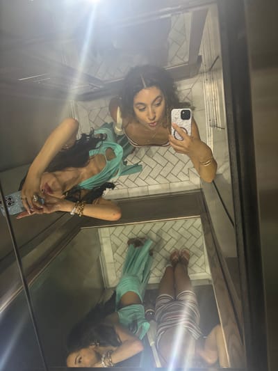 The image shows a reflection of two women in a mirrored elevator. The woman in the foreground is taking a selfie with her phone, wearing a light blue dress and jewelry. The second woman is in the background, wearing a striped skirt and sandals. The elevator walls are metallic, and the floor is tiled. The image appears to be taken in San Juan, Puerto Rico, a city in the United States.